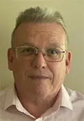 A middle-aged man with short grey hair, glasses, and a light-coloured collared shirt is facing the camera and smiling slightly against a plain background.