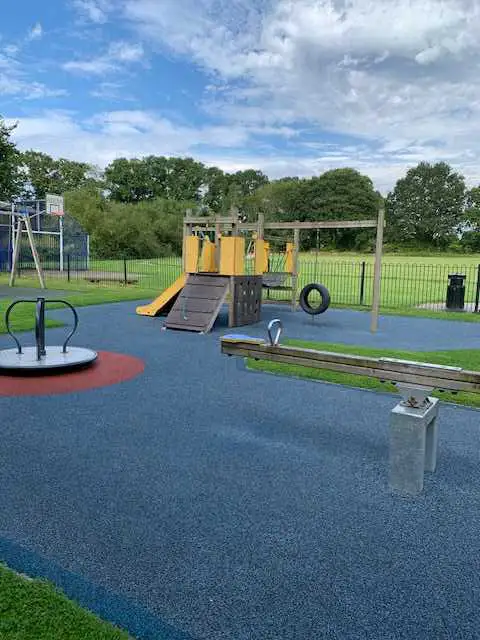 Children’s playground with a yellow climbing frame and slide, a seesaw, a roundabout, and blue safety flooring. The area is surrounded by green grass, trees, and a black fence under a partly cloudy sky.