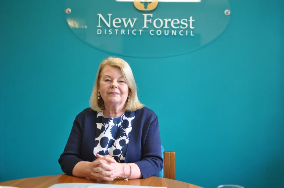 A woman with blonde hair, wearing a navy blazer and patterned blouse, sits at a table in front of a sign that reads New Forest District Council on a teal wall.