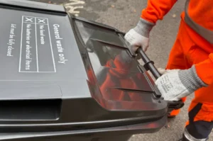 A person wearing orange high-visibility clothing and gloves is pushing a black general waste bin with a closed lid on a street. The bin has clear labelling for waste restrictions.