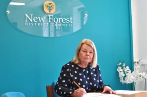 A woman with blonde hair and glasses sits at a desk, writing on papers, in front of a teal wall with a sign that reads New Forest District Council. White flowers are on the desk beside her.