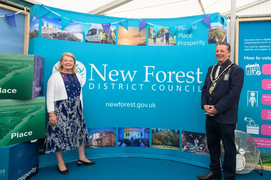 Two officials stand smiling in front of a blue New Forest District Council display, with images of nature and local places. They are indoors, beside large blocks labelled “Place” and a small electric fan.
