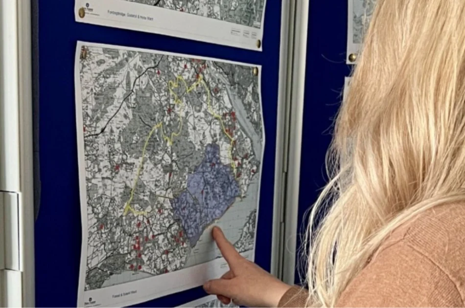 A person with long blonde hair points at a black-and-white map with coloured markings displayed on a wall among other maps.