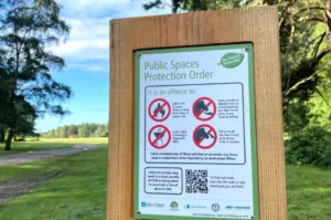 A wooden sign displays rules for a Public Spaces Protection Order in a grassy park, listing offences like lighting barbecues and feeding ponies. Blue sky and trees are visible in the background under a sunny sky.