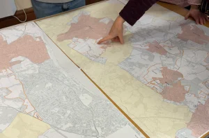 A person points to a specific area on a large, detailed map laid out on a table, while another person stands nearby. The map includes several outlined regions in different colours.